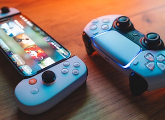 A gaming scenario featuring a smartphone with a game interface next to two controllers on a wooden surface. The smartphone displays vibrant images and icons, while the controllers feature a sleek, modern design with buttons and joysticks prominently visible. Warm lighting enhances the scene.