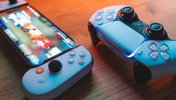 A gaming scenario featuring a smartphone with a game interface next to two controllers on a wooden surface. The smartphone displays vibrant images and icons, while the controllers feature a sleek, modern design with buttons and joysticks prominently visible. Warm lighting enhances the scene.