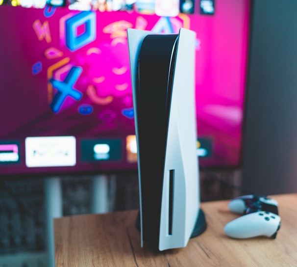 A gaming console is placed on a wooden table with a game controller nearby. In the background, a television screen displays colorful graphics, including geometric shapes and vibrant colors.